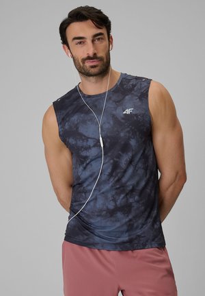 Mężczyzna w bezrękawnikowej, granatowej koszulce sportowej w stylu tie-dye i różowych szortach, słuchający muzyki na białych słuchawkach na jednolitym tle.