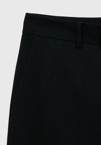 Dettaglio della vita di pantaloni neri con passante per cintura e tasca laterale su uno sfondo chiaro e neutro.