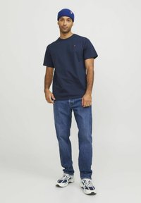 T-shirt di cotone blu navy con collo rotondo, maniche corte e taschino sul petto, abbinato a jeans in denim blu e scarpe da ginnastica bianche.
