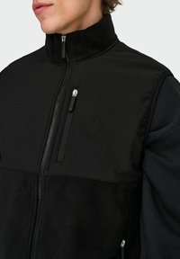 Uomo che indossa una giacca in pile nera con cerniera frontale e tasca sul petto con cerniera, che mostra il torso superiore e il viso nella parte inferiore in primo piano.