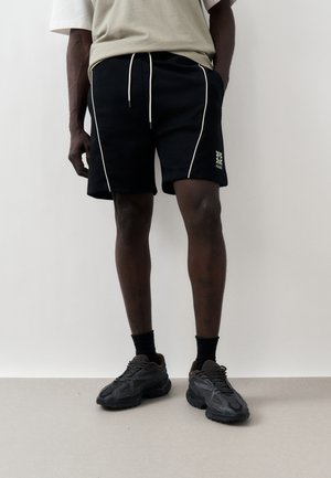 Mand iført sorte snørre-short med hvid piping og logo, sorte sokker og sorte chunky sneakers, stående mod en ensfarvet hvid væg.