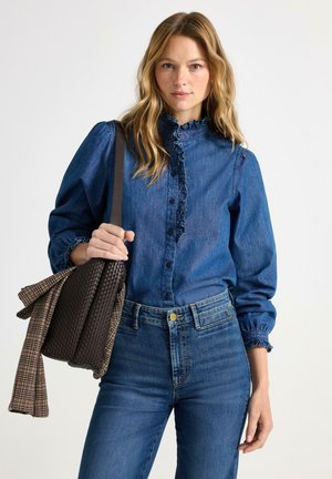 Denimblus i mörkblå med volangkrage, knäppning fram och puffärmar. Kombinerad med högmidjade, medelblå jeans och en brun väska.