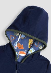 Tumesinise kapuutsiga, millel on roheline äär ja mustriline sinine vooder, kus on kujutatud loomi ja navigatsioonimärke. Pehme puuvillane materjal. Ees lukuga.