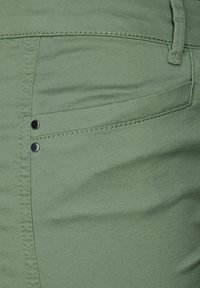 Nära bild av en grön byxficka med dubbel söm, två metallnit och detalj av bältesöglan.