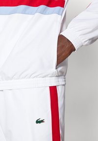 Λευκό αθλητικό μπουφάν με κόκκινες και ανοιχτό μπλε οριζόντιες ρίγες, ελαστικές μανσέτες, τσεπάκι με φερμουάρ και μικρό πράσινο λογότυπο κροκόδειλου στο πλάι.