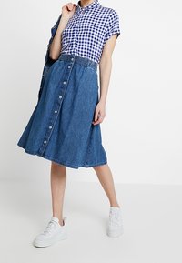Chemise à carreaux de type boutonnière en bleu et blanc, associée à une jupe en denim avec une taille smockée et une fermeture à boutons sur le devant. Baskets blanches.