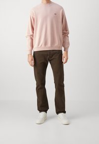 Sudadera rosa claro con cuello y puños acanalados, combinada con jeans marrón oscuro y zapatillas blancas, de pie frente a un fondo liso.