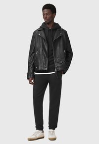 Chaqueta de motorista de cuero negro con herrajes plateados, superpuesta sobre una sudadera negra y una camiseta blanca, combinada con pantalones de chándal negros y zapatillas blancas.