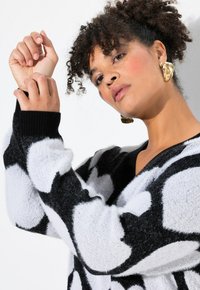 Zwart-wit gestreept sweater met een V-hals, gebreide textuur en zwarte ribbels bij de mouwen. Model draagt gouden oorbellen en heeft krullend haar.