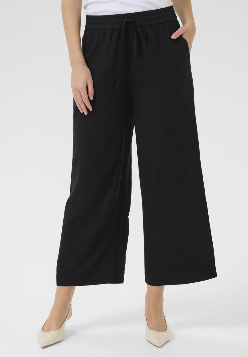 Personne portant un pantalon noir à jambes larges avec taille à cordon et poches, associé à des talons blancs à bouts pointus, debout sur un fond neutre.