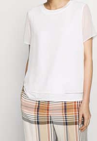 Haut blanc à manches courtes en tissu lisse avec des manches transparentes. Associé à un pantalon à carreaux avec des rayures orange, marron et noires.