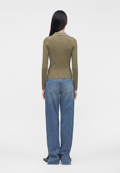 Femme aux longs cheveux foncés, debout de dos, portant un haut côtelé à manches longues vert olive et un jean bleu ample avec poches arrière.