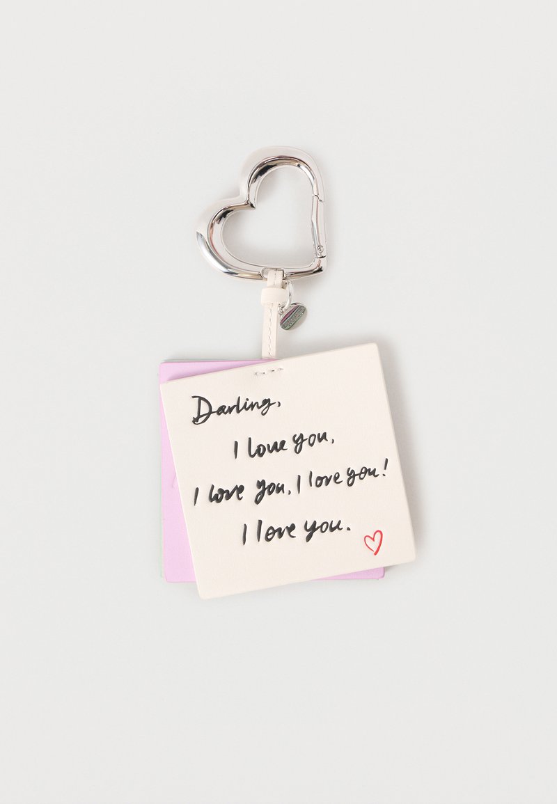 Ασημένιο κούμπωμα σε σχήμα καρδιάς προσαρτημένο σε ένα κρεμ και ροζ κάρτα με χειρόγραφο κείμενο που εκφράζει αγάπη, με κόκκινο διακοσμητικό καρδιάς.