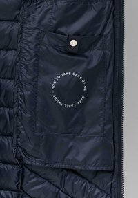 Interno di un giubbotto puffer blu navy con una tasca con zip e bottone a pressione, che mostra le istruzioni per la cura in testo circolare attorno ad essa.