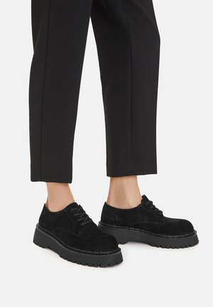 Zapatos de ante negro con cordones, suela gruesa y texturizada y detalle de costuras, combinados con pantalones negros de corte entallado.