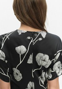 Kobieta z brązowymi włosami, prezentująca tył czarnej bluzki z białym kwiatowym wzorem oraz małym zapięciem przy dekolcie.