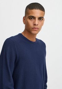 Marineblå teksturert strikkegenser med rund hals, lange ermer og en tettsittende silhuett. Glatt stoff med et subtilt mønster.