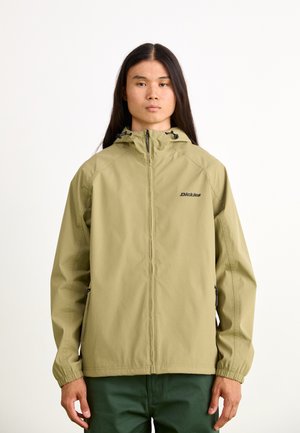 Chaqueta impermeable de un verde oliva claro con capucha, cierre frontal con cremallera y puños elásticos. Presenta un logo en el pecho y bolsillos laterales.