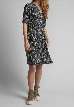 Femme portant une robe noire à manches courtes avec un imprimé floral blanc, des boutons en bois et des bottines marron à lacets, sur un fond uni.