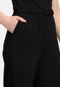 Zwarte jumpsuit met een aansluitende taille en een riem, zijzak en een zachte, matte stoftextuur.