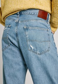 Svetlo modri traper hlače z ohlapnim krojem, ki imajo dva zadnja žepa, rjav usnjen trak na pasu in obrabljene detajle.