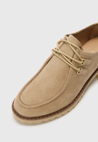 Beige Wildlederschuh mit runder Zehe, brauner Gummisohle und dekorativen goldenen Ösen. Verfügt über hellfarbene Schnürsenkel und Details in der Naht.