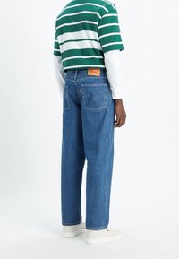 Hombre vestido con jeans rectos azules, camisa de manga corta a rayas verdes y blancas sobre una camisa de manga larga blanca, y zapatillas blancas, de pie.
