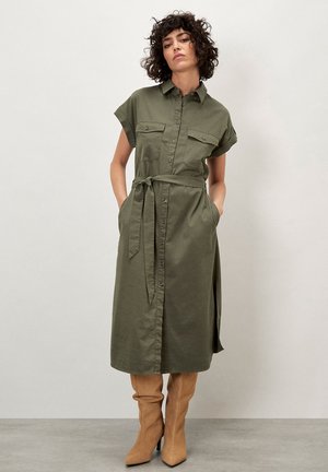 Olive green shirt dress with short sleeves, button front, two chest pockets, and a tie waist. Worn with tan knee-high fitted boots.