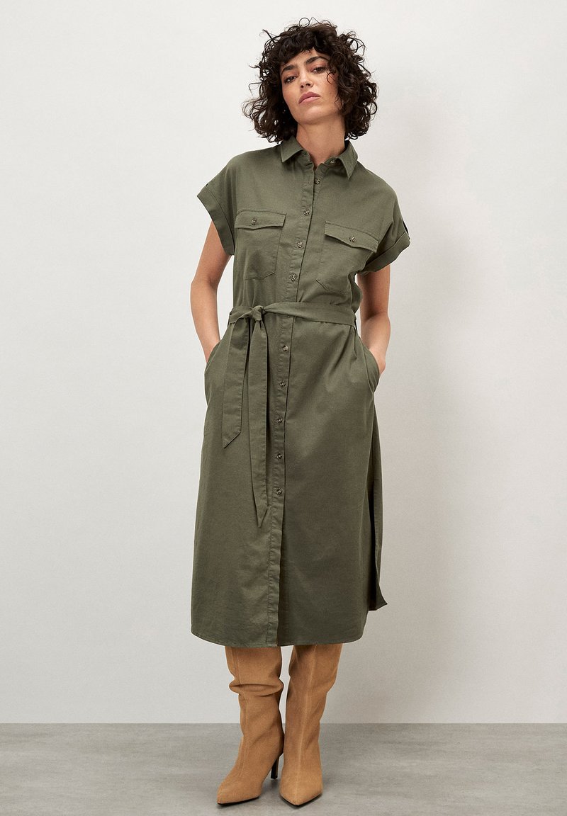Olivgrünes Hemdblusenkleid mit kurzen Ärmeln, Knopfleiste, zwei Brusttaschen und einem Bindegürtel in der Taille. Getragen mit beigen knielangen, enganliegenden Stiefeln.