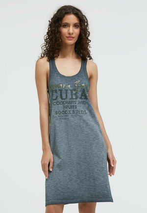 Frau mit lockigem Haar, die ein ärmelloses graues Kleid mit dem Schriftzug „Havana Bay Cuba“ trägt, in einer lässigen Pose vor hellem, einfarbigem Hintergrund.