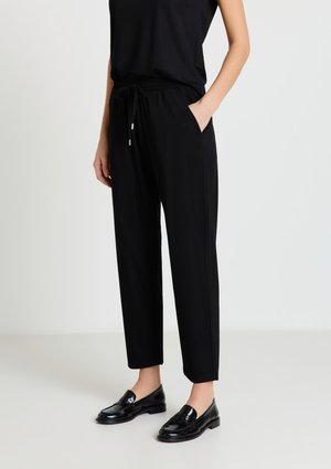 Femme portant un pantalon noir effilé avec une ceinture élastique et un cordon de serrage, associé à des mocassins en cuir noir, debout devant un fond uni.