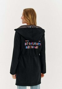 Sieviete ar blondiem matiem, ģērbta melnā kapuces jakā ar krāsainu tekstu "Lai tavi sapņi vada ceļu" uz muguras, apvienota ar gaiši ziliem džinsiem.