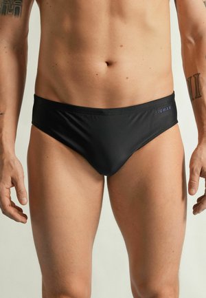 Mannelijke torso met zwarte zwemslips met een klein logo op de linkerheup tegen een effen lichte achtergrond.