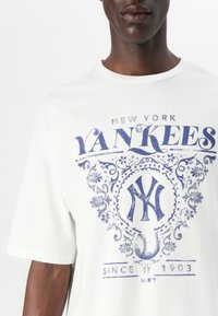 Baltas medvilnės marškinėliai su mėlynai atspausdintu "New York Yankees" logotipu su puošniomis detalėmis ir tekstu "Nuo 1903", apvaliu iškirpimu, trumpomis rankovėmis.