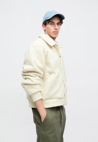 Φλις μπουφάν σε κρεμ απόχρωση με γιακά, κούμπωμα με τρουκς και πλαϊνές τσέπες. Φορώντας το με παντελόνι χακί και ανοιχτό μπλε καπέλο.