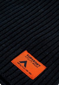 Tissu en maille côtelée noire avec une étiquette orange affichant "SUPERDRY UTILITY WEAR" et un logo triangulaire ; la texture semble douce et chaude.