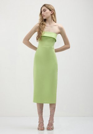 Donna in piedi con le mani dietro la schiena, che indossa un abito midi senza spalline di colore verde lime e sandali beige con lacci, su uno sfondo neutro.
