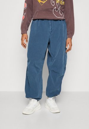 Person iført løse, blå snorebukser, hvide sneakers og en brun grafisk sweatshirt, stående mod en ensfarvet baggrund.