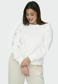 Felpa bianca con maniche a palloncino e dettagli floreali ritagliati. Design semplice e texture morbida, abbinata a pantaloni chiari.