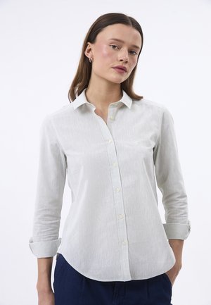 Jeune femme portant une chemise blanche boutonnée avec manches retroussées et pantalon bleu marine, debout devant un fond blanc uni.