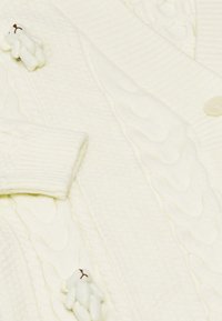 Valkoinen cable-knit neuletakki, jossa on kaksi pientä valkoista nallekarhuhakemusta kiinnitettynä nappien ja hihansuiden lähelle.