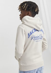 Felpa beige con stampa blu che recita "JACK & JONES Denim Crew Collective" sulla schiena. Presenta un cappuccio con coulisse e polsini a coste.