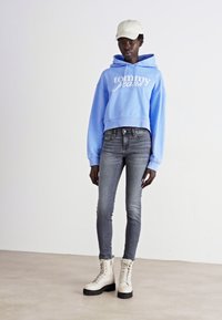 Klippt ljusblå hoodie med vitt logotyp, grå tajta jeans och krämfärgade kängor med tjocka svarta sulor. Ljus basebollkeps fulländar outfiten.