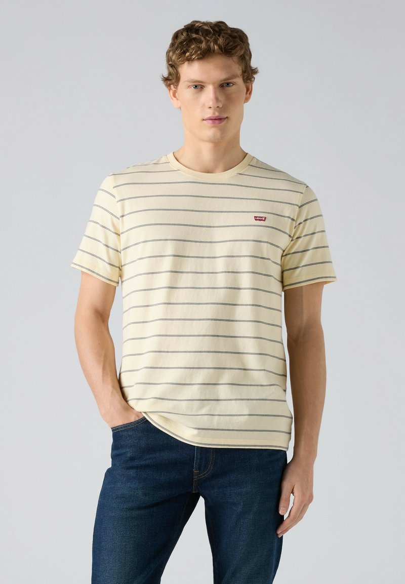 Jeune homme aux cheveux bouclés portant un t-shirt Levi's jaune clair rayé et un jean bleu foncé, debout avec une main dans la poche.