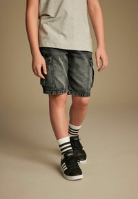 Pantalones cortos de mezclilla negros tipo cargo con múltiples bolsillos, combinados con zapatillas negras y calcetines a rayas blancas. Camiseta gris de cuello redondo parcialmente visible.