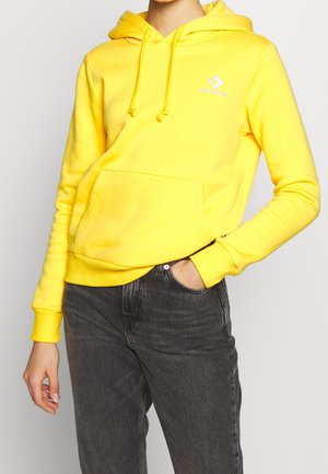 Persona con sudadera con capucha Converse de color amarillo brillante y vaqueros negros, con una mano en el bolsillo y la otra relajada a un lado.