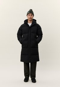 Vīrietis, kurš valkā garu melnu polsterētu mēteli ar kapuci, melnu cepuri, tumšas bikses un melnus apavus, stāv pret vienkāršu gaišu fonu.