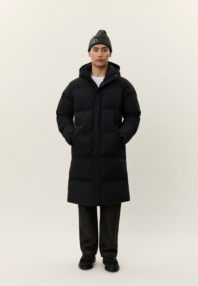 Vīrietis, kurš valkā garu melnu polsterētu mēteli ar kapuci, melnu cepuri, tumšas bikses un melnus apavus, stāv pret vienkāršu gaišu fonu.