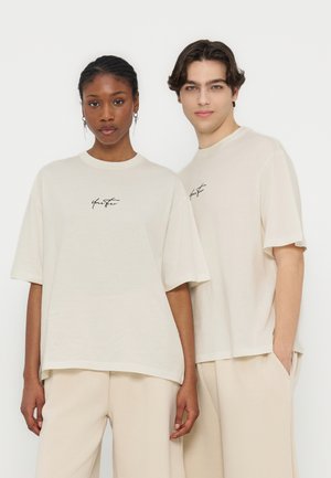 Dwa modele noszą pasujące luźne beżowe koszulki z małym czarnym tekstem pisanym ręcznie, zestawione z jasnobeżowymi spodniami na jednolitym białym tle.