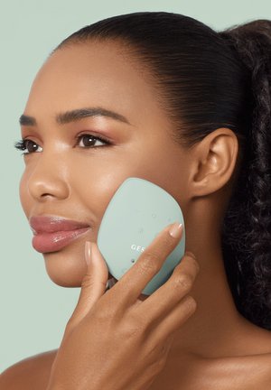 Vrouw met een gladde huid houdt een muntgroen gezichtsapparaat met knoppen tegen haar rechterwang, waarmee ze het gebruik voor huidverzorging demonstreert.